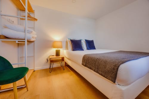 Quarto iluminado nas Arrabia Guest Houses com cama queen, almofadas azuis, criado em madeira, luminária, poltrona verde e piso de madeira