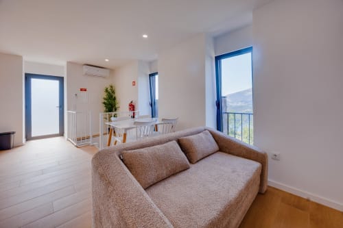 Quarto de hóspedes moderno e luminoso no Arrabia Glamping com sofá bege, mesa de jantar e vista para rio e montanhas através de janela de vidro azul