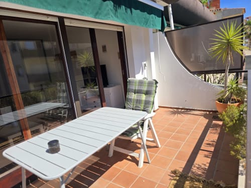 Terrasse ensoleillée avec table et chaise blanches, plantes palmiers en pots et portes vitrées donnant sur un appartement à Roses