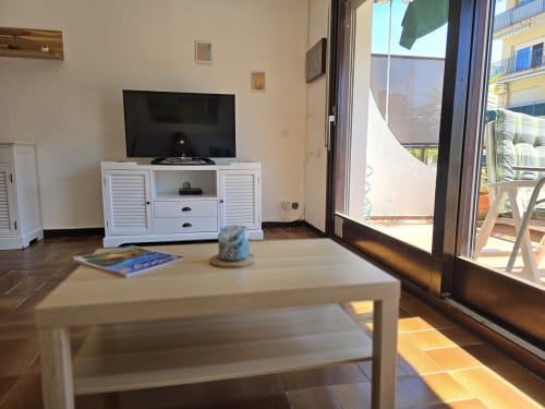 Salon douillet d'un studio avec meuble TV blanc, table et portes coulissantes donnant sur la terrasse