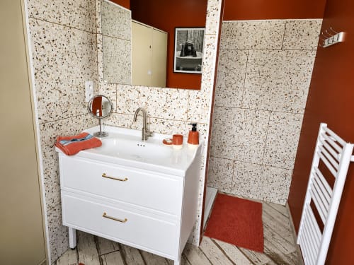 Salle de bains moderne avec vanité blanche, carrelage en terrazzo et accents rouge rouille à la villa Ô de Mer à Sangatte