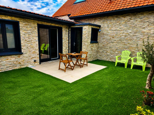 Patio du jardin de la Villa Ô Gris-Nez avec gazon synthétique, table en bois et chaises vert vif face à la maison en briques près de la plage
