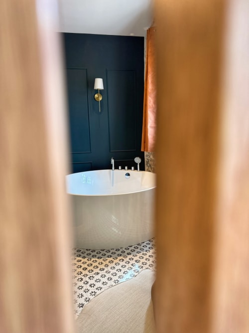 Salle de bain moderne avec baignoire blanche indépendante, murs bleu canard, carrelage à motifs et appliques en laiton à la Bastide