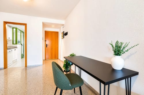 Entrada de apartamento moderno en Valencia con escritorio negro, silla verde, jarrón blanco con plantas, cocina y puerta naranja visible