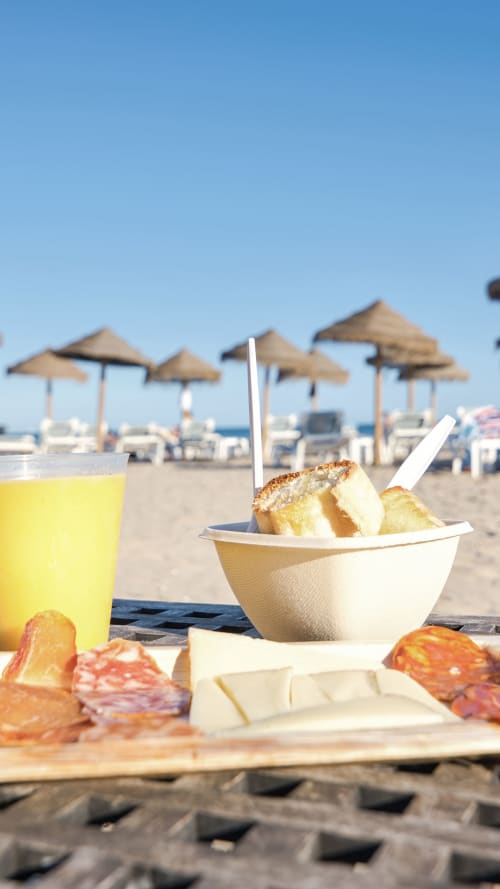 Picnic en la playa con zumos, pasteles y embutidos en apartamento de Valencia junto al mar con sombrillas de fondo