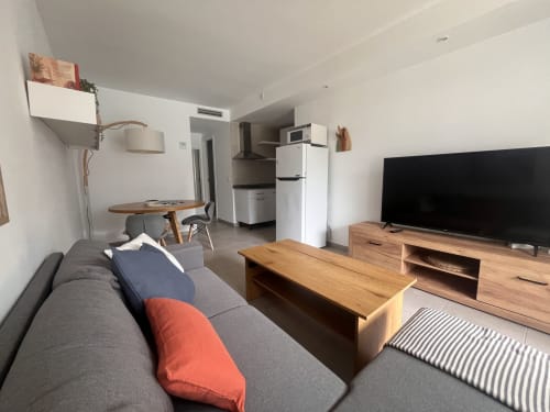 Moderno salón con sofá gris, mueble TV de madera, cocina americana y zona de comedor en Corralejo