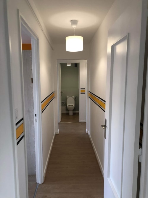 Couloir lumineux de l'hôtel Mattbnb Metz avec murs blancs, parquet, bandes jaunes et salle de bains commune