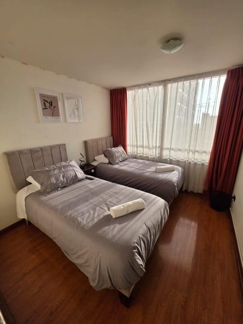 Camas gemelas con ropa de cama gris en luminosa habitación del Your Place Spa, Antofagasta, con amplios ventanales y cortinas rojas