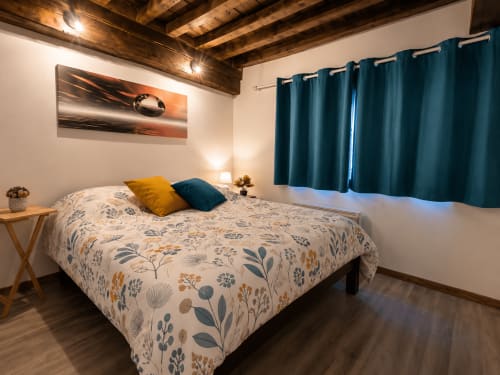 Chambre cosy à l'appartement Systeo à Châtillon-sur-Chalaronne avec literie florale, rideaux bleu-vert et poutres apparentes