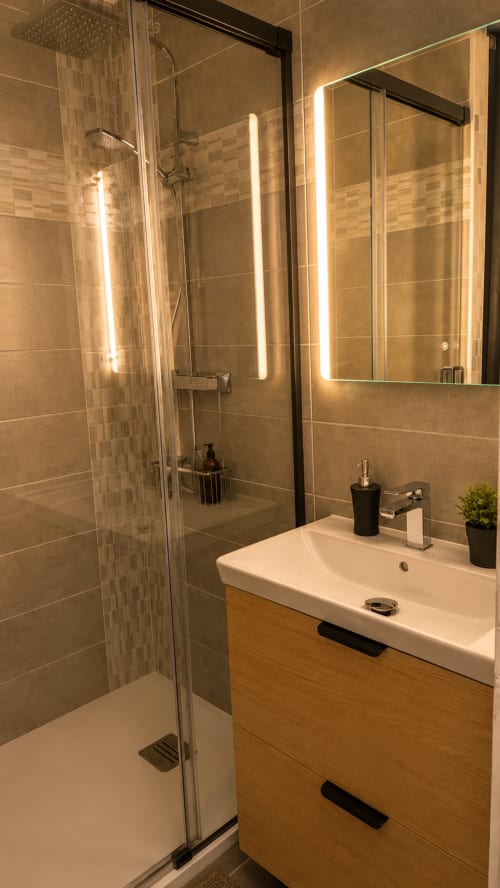 Salle de bains moderne de l'appartement Systeo à Mâcon avec miroir rétroéclairé, meuble-lavabo en bois et douche à l'italienne