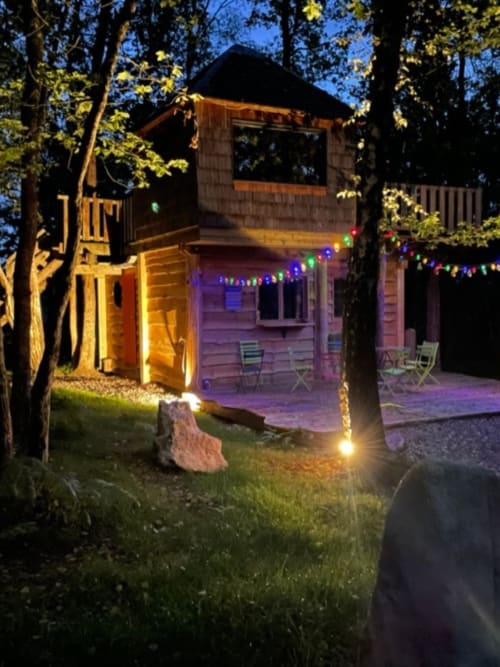 Illuminated treehouse cabin at Les Murmures du Bois with colorful string lights and seating area at dusk