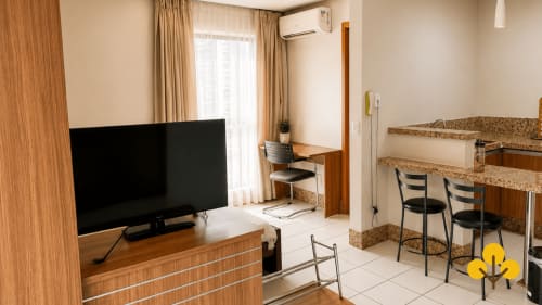 Sala de estar moderna com TV, bancada de cozinha, mesa de trabalho e vista para a cidade em hotel de Goiânia