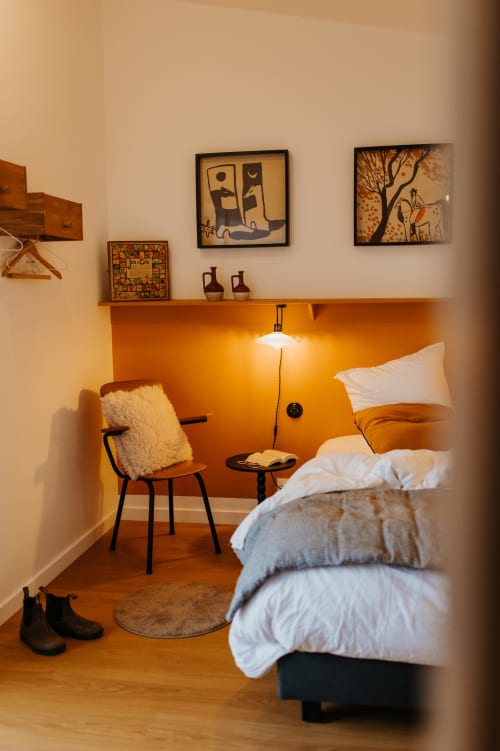 Chambre douillette aux Écuries du bout du monde à Landunvez : mur orange, lit confortable, fauteuil de lecture et œuvres encadrées.