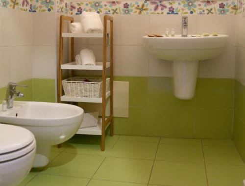Bagno luminoso con piastrelle verde lime, sanitari bianchi, mensola in legno con articoli da toilette, lavabo con piedistallo e bidet