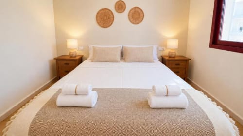 Moderno dormitorio en apartamento Aloha Calpe con ropa de cama blanca, mesillas de madera, toallas enrolladas y decoración tejida en la pared