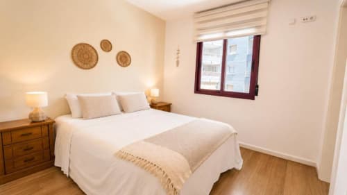 Luminoso dormitorio en apartamento Aloha Calpe con cama blanca, mesillas de madera, arte textil y vistas al mar