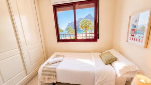 Dormitorio luminoso en Aloha Calpe con vistas al mar, ventanas hacia montañas y palmeras, cama blanca y comodidades modernas