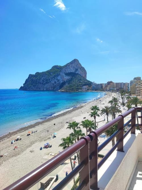 Vista de balcón de la playa de Calpe con la formación rocosa Peñyal d'Ifach, arena, palmeras y mar Mediterráneo