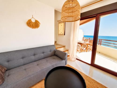 Moderno salón en dúplex Aloha Calpe con sofá gris, lámpara colgante tejida y vistas al mar desde las puertas del balcón