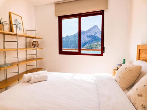 Moderno dormitorio en Aloha Calpe con vistas al mar y picos montañosos a través de ventana con marco rojo, estantes de madera y ropa de cama blanca