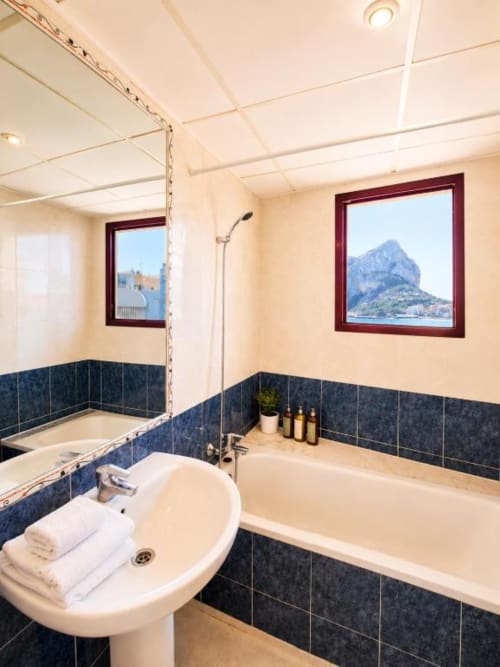 Baño moderno con ventana con vista al mar en apartamento dúplex Aloha Calpe, con bañera, lavamanos y detalles en azulejos oscuros