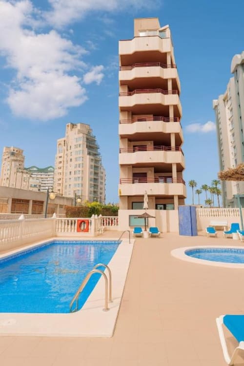 Edificio de apartamentos Aloha Calpe con piscina, tumbonas y palmeras bajo cielo azul