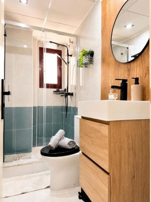 Baño moderno en apartamento Aloha Calpe con vanidad de madera, espejo redondo, ducha de cristal con azulejos turquesa y estilo costero
