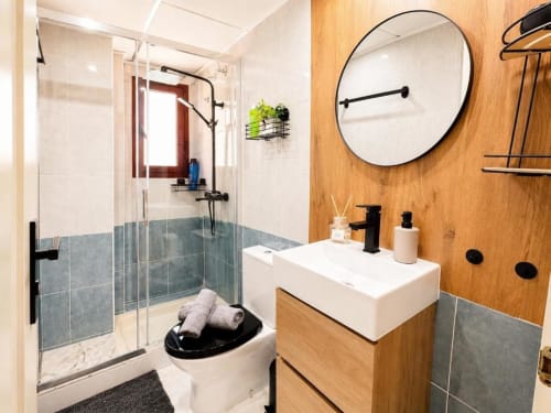 Baño moderno en apartamento Aloha Calpe con pared de madera, ducha abierta, vanidor blanco y espejo redondo