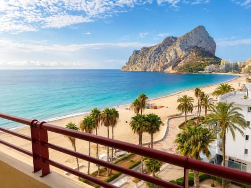 Vista del balcón hacia la playa de Calpe con la formación rocosa Peñón de Ifach, palmeras y el Mar Mediterráneo desde el apartamento Aloha Calpe