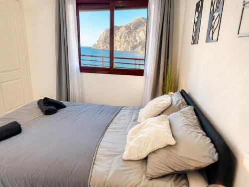 Habitación con vistas al mar de los acantilados de Calpe desde el balcón, cama con almohadas crema, ropa de cama gris, apartamento moderno