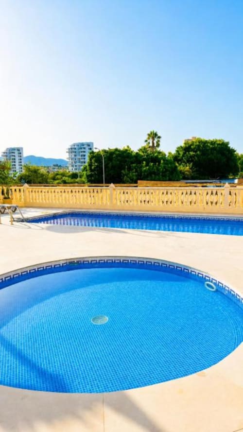 Complejo residencial Aloha Calpe con dos piscinas azules con barandillas amarillas, palmeras y vistas a la montaña