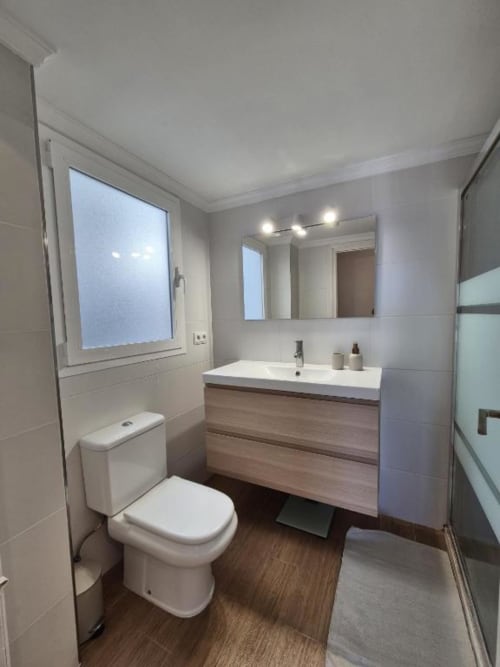 Baño moderno en apartamento del Casco Antiguo de Aloha Calpe con lavabo blanco, tocador de madera, inodoro, espejo y ventana con vistas al mar