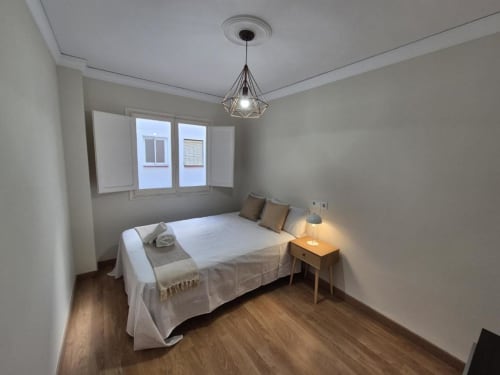 Dormitorio luminoso en apartamento del Casco Antiguo, Aloha Calpe, con cama de sábanas blancas, mesita de madera, luz colgante y vistas al mar
