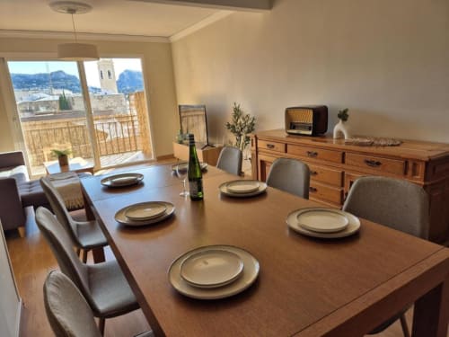 Elegante comedor en apartamento de Calpe con vistas al mar, mesa de madera dispuesta para comer y vistas a la montaña a través de puertas de cristal
