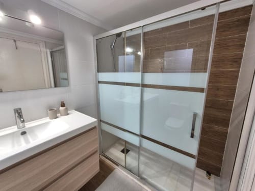 Baño moderno en apartamento Aloha Calpe con ducha de cristal, lavabo blanco y detalles de madera y azulejos