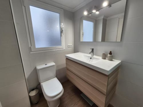 Baño moderno en apartamento del Casco Antiguo del hotel Aloha Calpe con lavabo blanco, tocador de madera, inodoro, espejo y ventana esmerilada