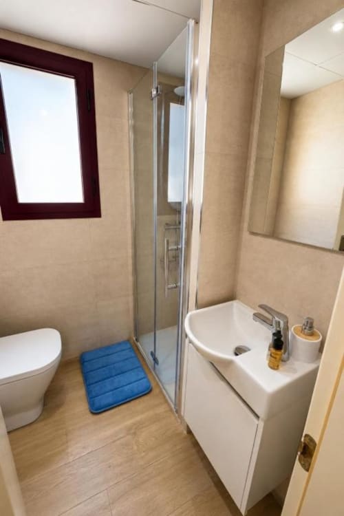 Baño moderno en Aloha Calpe Dreams Apartment con ducha, lavabo, inodoro y tapete azul sobre piso de madera.