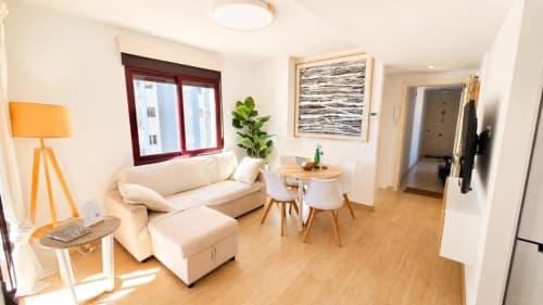 Luminoso salón del apartamento Dreams en Aloha Calpe con sofá crema, suelos de madera y vistas al mar desde marco granate