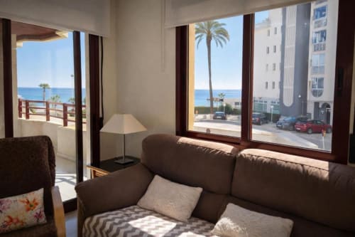 Sala de estar del apartamento Sea Palm en Calpe con sofá marrón, vistas al mar desde las puertas del balcón, palmeras y costa mediterránea