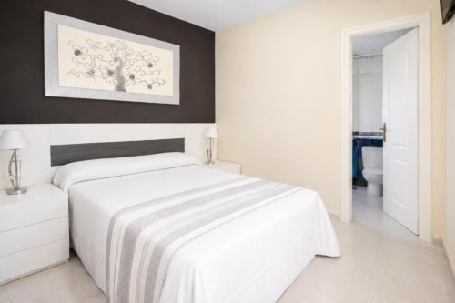 Dormitorio moderno en Aloha Calpe Sea Palm Apartment con ropa de cama blanca, cabecero oscuro y acceso a baño privado