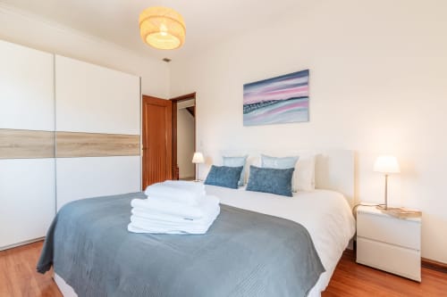 Quarto luminoso com cama de casal, almofadas azuis, roupa de cama branca, guarda-roupa em madeira e quadros com vista de jardim em Arestas e Planetas