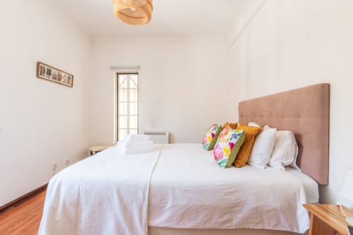 Quarto luminoso com cabeceira estofada em rosa, roupa de cama branca, almofadas coloridas, piso de madeira e vista para jardim