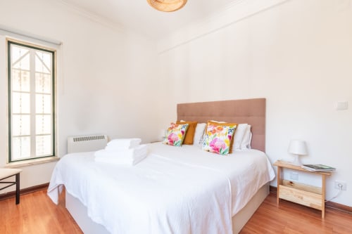 Quarto luminoso com cabeceira bege, roupa de cama branca, almofadas coloridas, criado em madeira e vista para o jardim em Arestas e