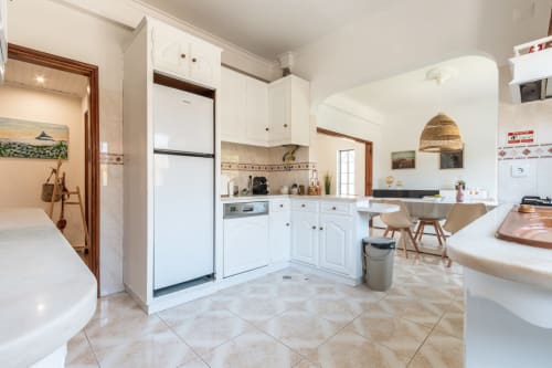 Cozinha aberta integrada à sala de jantar da villa Amora, com armários brancos, eletrodomésticos em aço inoxidável e piso em terracota