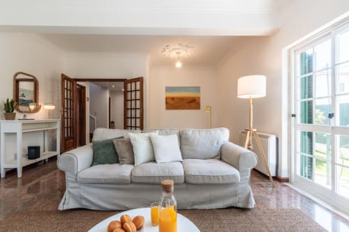 Sala de estar luminosa na villa Amora com sofá bege, vista para o jardim através de portas francesas, lareira e mobiliário moderno