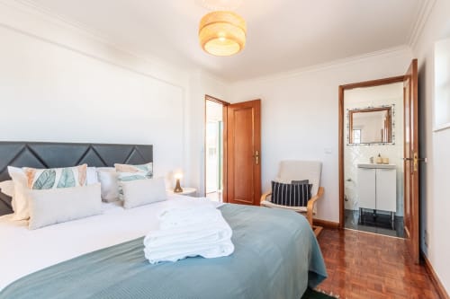 Quarto luminoso com cama queen, cabeceira cinza, portas de madeira, banheiro privativo e luminária de rattan na villa Amora