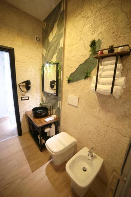 Bagno moderno delle Roma Imperial Suites con sanitari bianchi, mobile in legno, lavabo nero, piante verdi alle pareti e portasciugamani