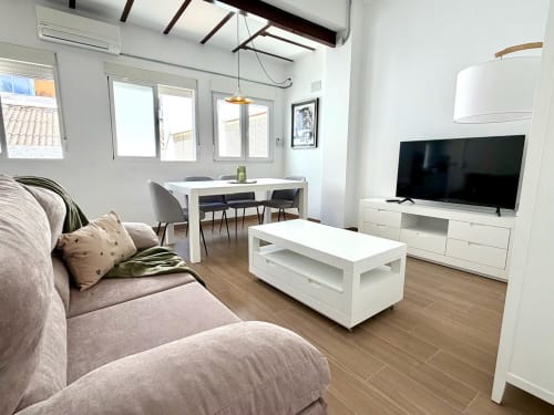 Salón moderno en loft de Valencia con sofá gris, televisor de pantalla plana, mesa de comedor blanca y ventanales luminosos con vistas a la ciudad