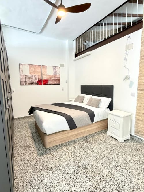 Dormitorio moderno en loft Stay Gest Valencia con cama, mesita de noche, ventilador de techo y escaleras a entresuelo