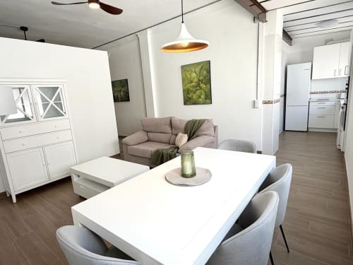 Moderno loft urbano en Valencia con mesa de comedor blanca, sillas grises, sofá topo, iluminación colgante y área de cocina